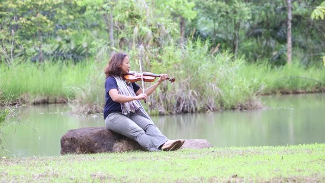 a asian woman with black hair, jean and scarf plays violin near the lake