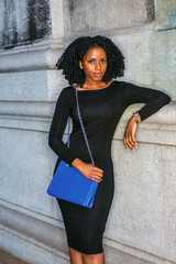 African American Woman street fashion in New York. Wearing long sleeve slim dress, wristwatch, carrying blue shoulder bag, young black girl with braid hairstyle standing against wall, looking at you..