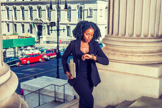 African American Business Woman Traveling, Working In New York. Holding Laptop Computer, Looking Down At Wristwatch, Young Black Lady With Braid Hairstyle Walking Into Office Building. Time Is Money.