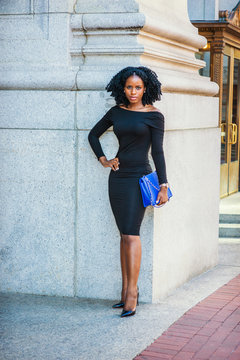 African American Business Woman working in New York. Wearing long sleeve, slim, off shoulder dress, carrying blue bag, young black lady with braid hairstyle standing outside office, greeting you..