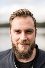 Laid back man with beard standing against a big lake looking at camera outdoors on a sunny day.