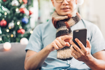Asia man wear santa hat use mobile call friends and sitting on sofa at Christmas tree in house,invite people to come winter festive holiday party.