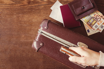 Tourist's hand holding old, leather, suitcase. Brown wallet and euro money with passport and tickets. Historical atmosphere with contemporary banknotes. Vintage style. Traveling or emigration concept.