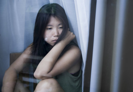 young sad and depressed Asian Chinese woman looking thoughtful through window glass suffering pain and depression in sadness concept