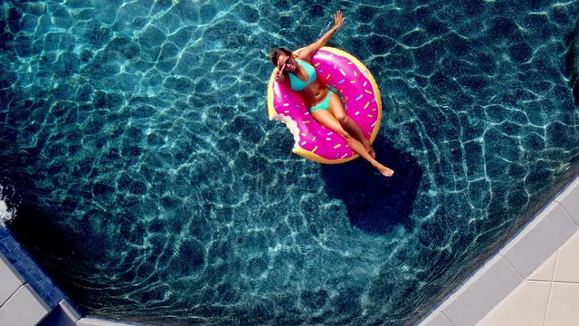 Young happy woman relaxing on inflatable pool toy in blue swimming pool on sunny day waiving at camera 