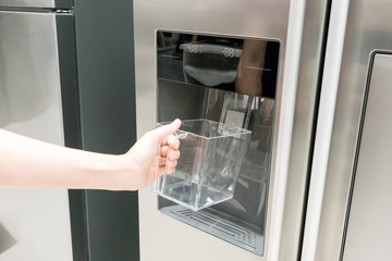 Closed up woman hand holding empty plastic container under the ice maker of refrigerator.