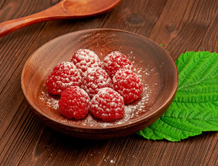 A dessert of ripe raspberries and powdered sugar on a wooden plate. Wooden spoon and a green leaf on wooden background.