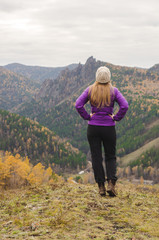 A girl in a lilac jacket looks out into the distance on a mountain, a view of the mountains and an autumnal forest by an overcast day. Free space for text
