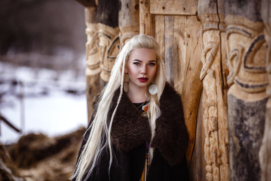 Outdoors portrait of beautiful furious scandinavian warrior ginger woman in a traditional clothes with fur collar, with sword in her hand and wooden Viking Village view on the background.