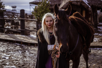 Obraz premium Outdoors portrait of beautiful furious scandinavian warrior ginger woman in a traditional clothes with fur collar, with sword in her hand and wooden Viking Village view on the background.