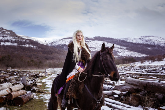Outdoors portrait of beautiful furious scandinavian warrior ginger woman in a traditional clothes with fur collar, with sword in her hand and wooden Viking Village view on the background.