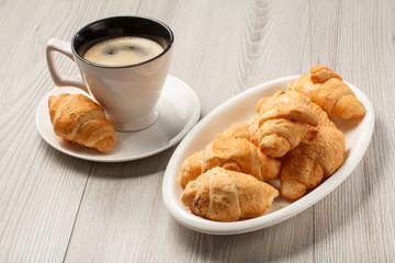 Cup of black coffee with saucer, fresh croissants on white porcelain dish