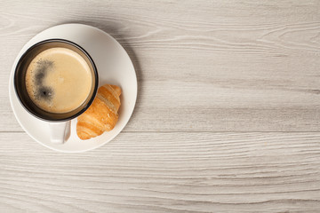 Top view of cup of black coffee with fresh croissant on saucer with copy space
