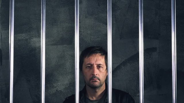Pensive man in jail behind the prison bars thinking about his criminal past and the mistakes he made, feeling guilt and remorse