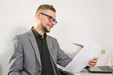 man with glasses and jacket smiles studying documents at table with laptop and printer at office