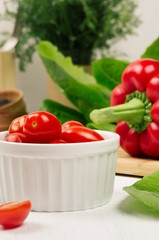Ripe red cherry tomato in white bowl with fresh leaves salad on white wood board, closeup.