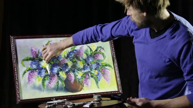 A man bearded artist in a blue sweater draws with palette knife painting flowers still life on easer in the studio