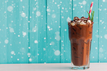 Homemade iced chocolate or cacao in glass topped with marshmallow and chocolate sauce on white wood table and blue background with snowfall. Concept to present bitter sweet and delicious drink.