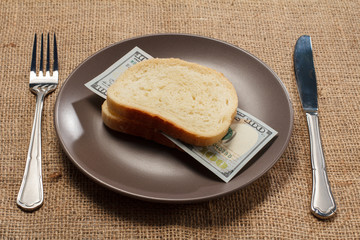 Two slices of bread with one hundred American dollars bill as hamburger of money on brown plate with fork and knife
