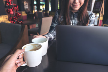 Close up image of two people clink white coffee mugs while working on laptop in cafe