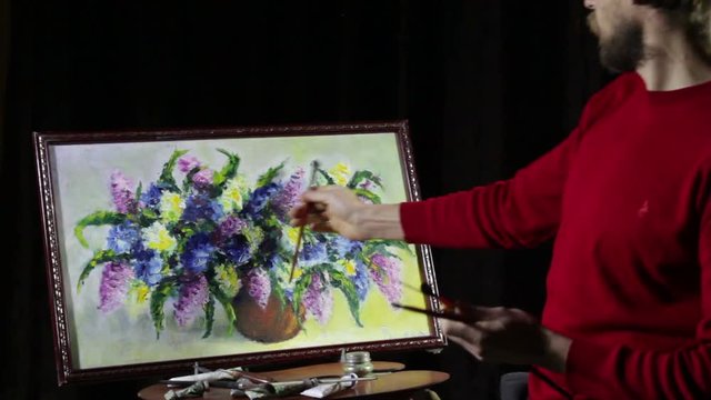 A man bearded artist in a red sweater draws an artistic brush painting flowers still life on easer in the studio
