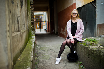 Fototapeta na wymiar Blonde girl at glasses and pink coat, black tunic and handbag posed at street.