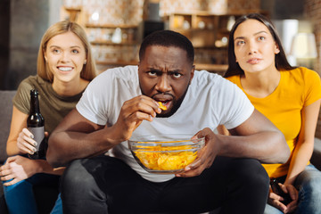 Snacks during game. Three glad jolly attractive friends looking up  while afro American man opening his mouth and eating chips