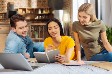  Group discussion.  Three pensive attractive young friends gazing  at the notebook while grinning and posing against blurred background 
