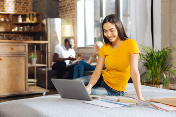 Diligent student. Appealing brunette young woman sitting on the bed while staring at the screen and smiling