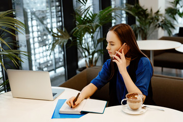 The young business woman works at office. The girl sits at a table, makes entries in the daily log and looks in the laptop.