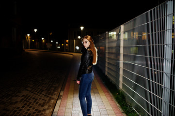 Night portrait of girl model wear on jeans and leather jacket against iron fence.