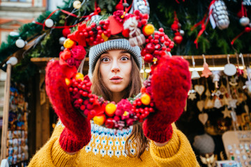 Portrait of surprised positive hipster woman looking through Christmas wreath over kiosk outdoors at christmas fair market on central square of the european city.