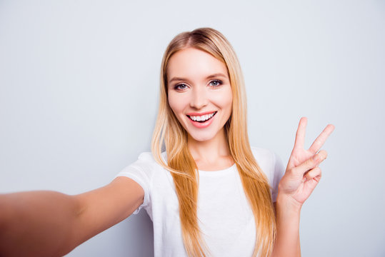 Cute charming lovely positive attractive young lady with blonde hair dressed in white t-shirt is taking a self portrait and demonstrating v-sign, isolated on grey background