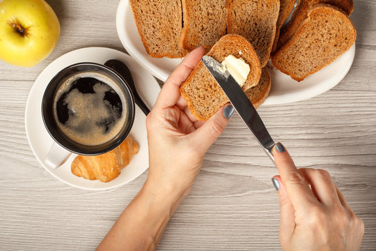View of female hands spreading butter on toast, cup of black coffee, croissant, bread and apple