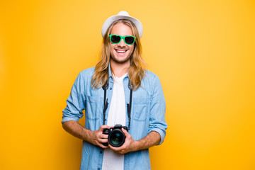 Give me  smile. Cheerful attractive young journalist in glasses, hat, casual outfit using photo camera over yellow background