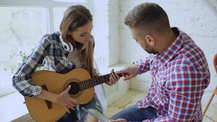 Young happy and loving couple study to play acoustic guitar using tablet computer and having fun while sitting at the table at home