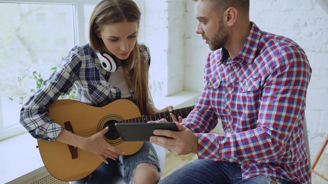 Young Happy And Loving Couple Study To Play Acoustic Guitar Using Tablet Computer And Having Fun While Sitting At The Table At Home