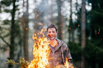 Happy funny crazy fearless man holding burning firewood in hands. Excited bizarre unusual boy laughing with stained dirty face in forest at nature. Fire in travel camp. Young male playing with flame.