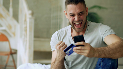 Attractive young man with smartphone and credit card shopping on the internet sit on bed at home