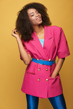 Beauty portrait of young african girl with afro hairstyle. Amazing girl wearing pink jacket, blue leggings and belt posing on yellow background. Studio shot.