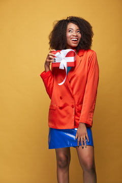 Colorful portrait of young african girl with afro hairstyle. Smiling girl wearing orange red jacket, blue latex skirt holds red present in her hand and posing on yellow background. Studio shot.