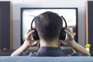 Back side of an Asian man wearing black heaphones in front of blurry out-of-focus television and...