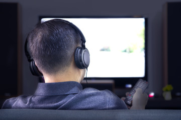 Back side of an Asian man wearing black headphones in front of blurry out-of-focus television and...