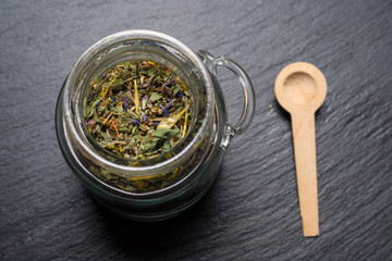 Herbs in glass jar with wooden spoon on black stone background
