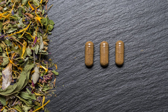 Herbal capsules and herbs on black stone background