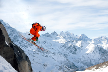 A professional skier makes a jump-drop from a high cliff against a blue sky leaving a trail of snow...