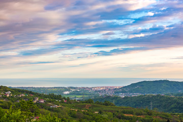 Fototapeta na wymiar View of the bay in the Black Sea from the height of the green mountains. Residential modern high-rise houses next to private cottages under beautiful clouds at dusk. Adler, Big Sochi, Russia.