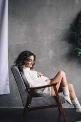 Caucasian pregnant woman with make up and curly hair in warm white sweater holds her belly, portrait of future mother, happy pregnancy, fashion portrait, stylish pregnant woman