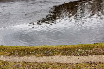 Frozen water at the shore of the pond with a path and lawn
