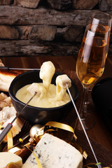 Gourmet Swiss fondue dinner on a winter evening with assorted cheeses on a board alongside a heated pot of cheese fondue with two forks dipping bread and white wine or champagne